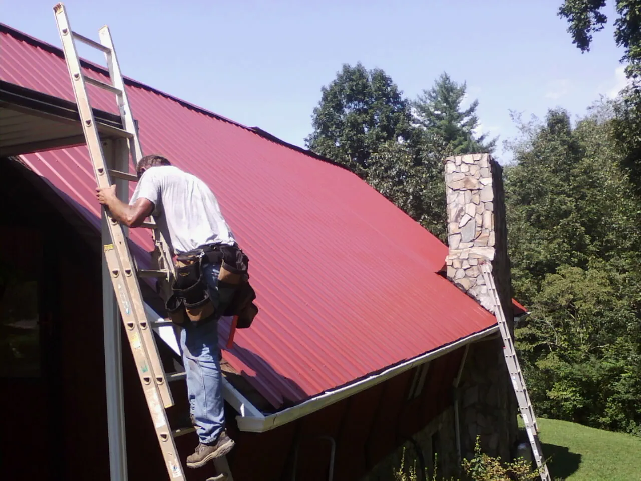 Roofer on a ladder beside a red metal roof and stone chimney — Soffit & Fascia Repair by Roofing Brothers in Heflin